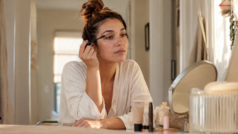 woman applying mascara at vanity mirror in morning light — 5 minute makeup routine for busy moms 2026